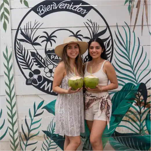 Dos mujeres posando con cocos frescos y bebidas tropicales frente al mural de bienvenida en Palapa Mar Azul, Playa de Monte Gordo.