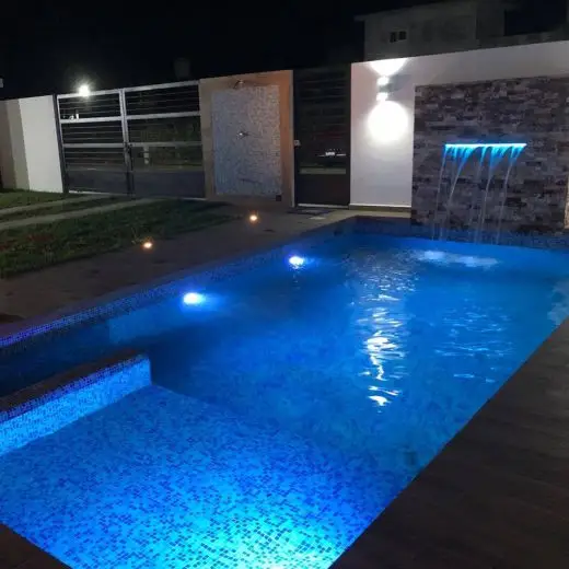 Alberca privada de lujo iluminada de noche en Villa Tortuga con luces LED azules, cascada sobre muro de piedra y área de poca profundidad.