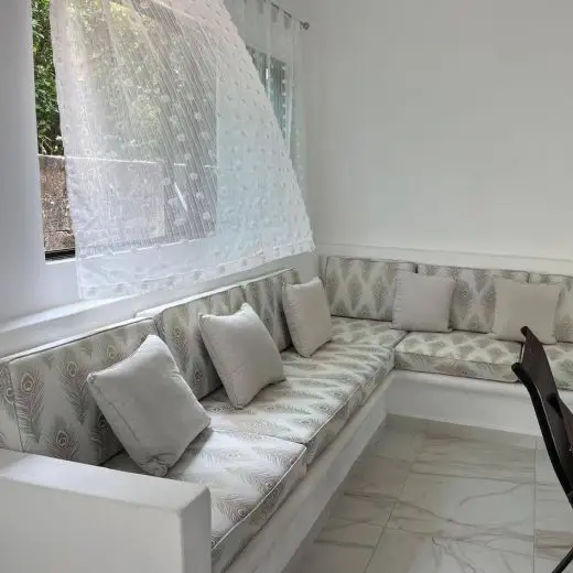 Luminosa y cómoda área común interior en Villa Tortuga con un moderno sofá esquinero integrado de diseño exclusivo, cojines y un gran ventanal con cortina de gasa ondeando, transmitiendo relajación y frescura cerca al mar. El suelo es de baldosa pulida.